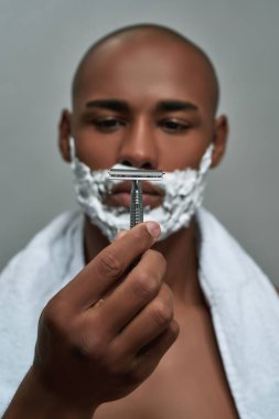 Yüzüne tıraş köpüğü sürmüş, elinde jiletle gri arka planda yalnız poz veren yakışıklı Afro-Amerikan bir adam.