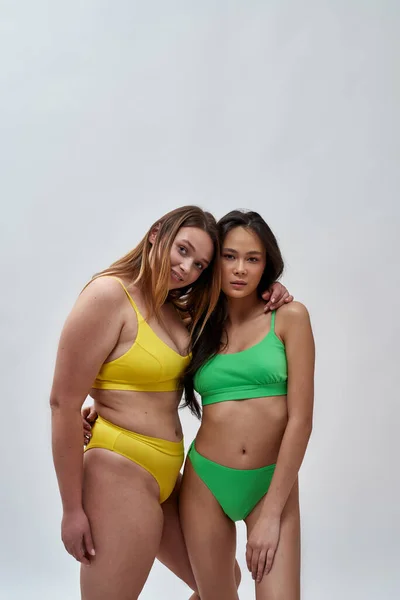 Deux belles jeunes femmes avec différentes formes de corps portant des sous-vêtements colorés regardant la caméra, embrassant tout en posant ensemble isolé sur fond de lumière — Photo