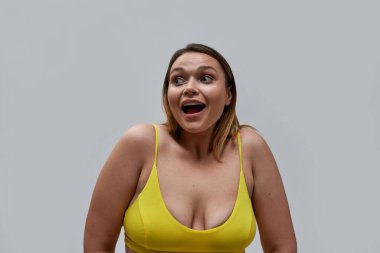 Gri arka planda izole edilmiş sarı sutyen giyen heyecanlı, kıvrımlı genç bir kadının portresi.