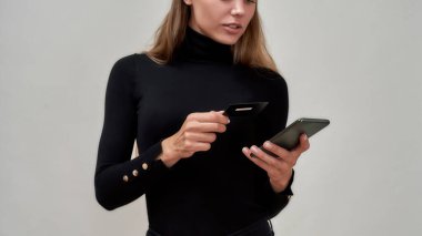 Siyah elbiseli, elinde kredi kartı olan, gri arka planda izole edilmiş bir şekilde poz verirken akıllı telefon kullanan beyaz bir kadının görüntüsü.