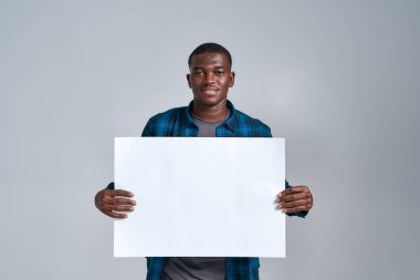 Neşeli, Afro-Amerikalı, günlük giysiler içinde, kameraya gülümseyen, boş afiş gösteren, önünde duran, gri arka planda izole edilmiş poz veren genç bir adam.