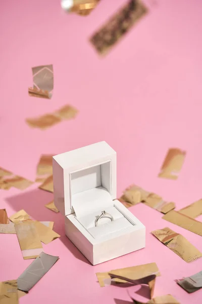 Romantic present for her. Vertical shot of elegant engagement diamond ring in white gift box and falling shiny golden confetti on pastel pink background