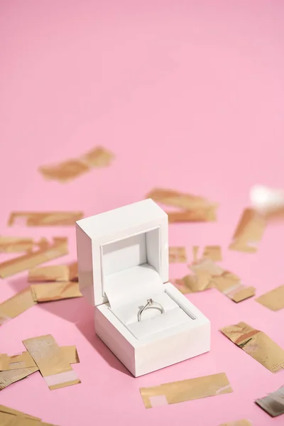 Marry me. Vertical shot of elegant engagement diamond ring in white gift box and shiny golden confetti on pastel pink background