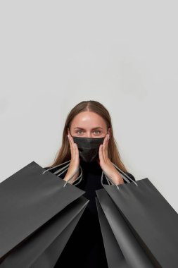 Siyah elbiseli ve koruyucu maskeli heyecanlı genç bir kadın kameraya bakıyor, her iki elinde de ışık altında izole edilmiş alışveriş torbalarıyla poz veriyor.