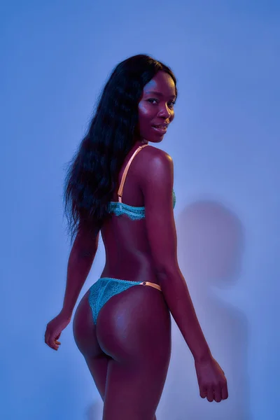 Sexy afro-américaine jeune modèle féminin avec un corps magnifique souriant tout en se tenant avec son dos à la caméra, posant en dentelle lingerie isolée en néon uv violet lumière bleue — Photo