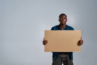 Ciddi bir Afro-Amerikan erkeği, günlük giysiler içinde, kameraya bakıyor, önünde boş afiş tutuyor, gri arka planda yalnız poz veriyor.