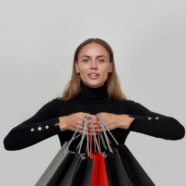 Siyah elbiseler giyen, kameraya gülümseyen ve alışveriş torbalarını gösteren, aydınlık arka planda yalnız poz veren genç beyaz bir kadın.