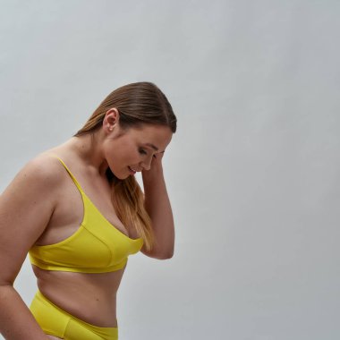 Gri arkaplanda izole edilmiş kamera için sarı iç çamaşırı giyen utangaç kadın modelin yan görüntüsü