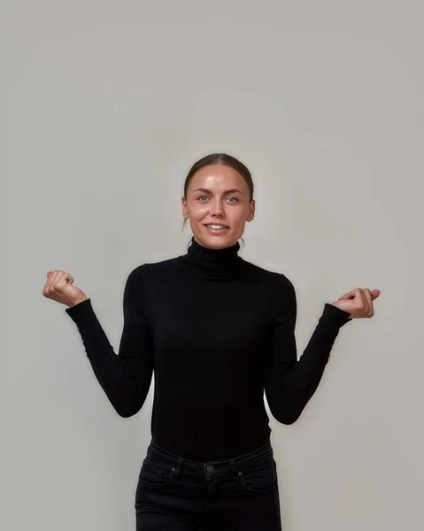 Mulher caucasiana encantada vestindo gola alta preta sorrindo para a câmera, de pé com as mãos levantadas apresentando seu produto isolado sobre fundo cinza — Fotografia de Stock