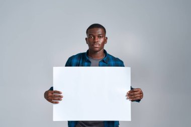 Ciddi bir Afro-Amerikan genci, günlük giysiler içinde kameraya bakıyor, boş afiş sergiliyor, önünde tutuyor, gri arka planda izole bir şekilde poz veriyor.