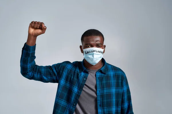 Grave chico afroamericano joven con máscara protectora con inscripción BLM mirando a la cámara, posando con el puño levantado aislado sobre fondo gris —  Fotos de Stock