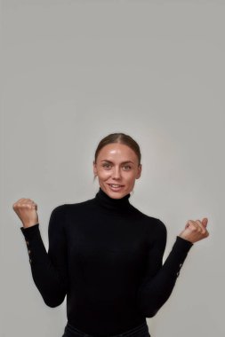 Siyah balıkçı yaka kazak giyen, kameraya gülümseyen, yumruklarını kaldırmış, gri arka planda izole olmuş beyaz bir kadın.