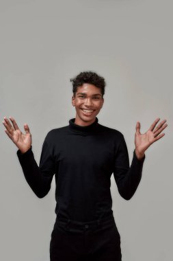 Heyecanlı genç adam kameraya zarif bir şekilde gülümserken elleri havada, gri arka planda yalnız poz veriyor.
