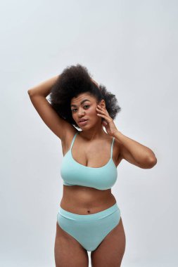 Kıvrımlı, genç, melez, afro saçlı, mavi iç çamaşırlı, kameraya poz veren, arka planda tek başına duran kadın model.