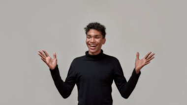 Siyah balıkçı yakalı mutlu genç adam gözleri kapalı gülerken elleri kaldırmış gri arka planda dikiliyor.