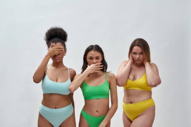 Farklı vücut şekillerine sahip çok ırklı genç kadınlar renkli iç çamaşırları giyiyorlar. Ağzı, gözleri ve kulakları kaplayan jestler gösteriyorlar. Hafif arka planda izole bir şekilde poz veriyorlar.