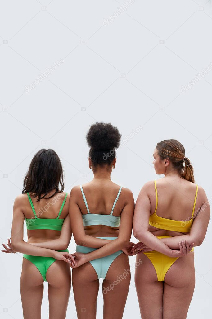 Rear view of three women with different body types in colorful underwear holding hands, supporting each other while posing isolated over white background