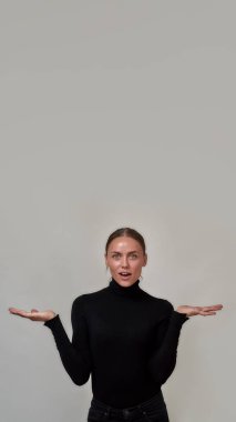 Siyah balıkçı yaka kazak giyen beyaz bir kadın çok heyecan verici ürününüzü sunuyor. Gri arka planda tek başına duruyor.