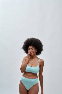 Afro saçlı, mavi iç çamaşırlı, beyaz arka planda yalnız poz verirken kameraya gülen neşeli, Afro Amerikalı bir kadın.