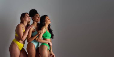 Farklı vücut şekillerine sahip üç farklı genç kadın renkli iç çamaşırları giyip yan yana gülümsüyor ve gri arka planda birlikte poz veriyorlar.