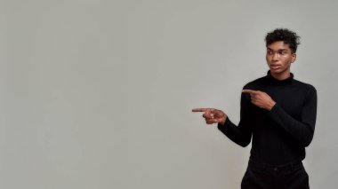 Siyah giyinmiş, piercing takmış, sol tarafı işaret eden, gri arka planda tek başına duran zarif bir genç.