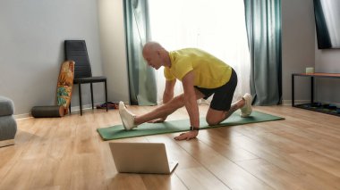 Yetişkin spor giyim eğitmeni evde online antrenman yapıyor, yoga minderinde egzersiz yapıyor, dizüstü bilgisayarda sanal fitness dersi alıyor.