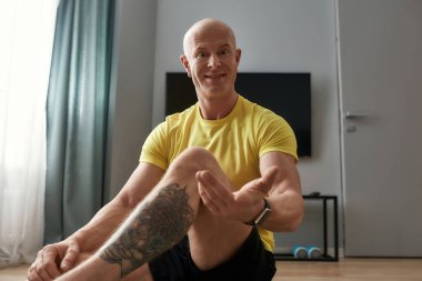 Evde yoga minderinde oturmuş, internetten eğitim alırken kameraya bakıp el kol hareketi yapan, sanal fitness dersi alan olgun bir antrenör.