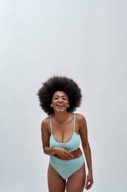 Heyecanlı, afro saçlı, mavi iç çamaşırı giyen, kameraya gülen, arka planda izole bir şekilde poz veren genç bir kadın.