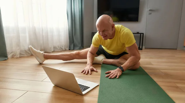 Спортивна зріла особиста фітнес-тренажер, що проводить онлайн-тренування, сидячи на йога килимку вдома і демонструючи спортивні вправи, маючи віртуальний фітнес-клас на ноутбуці — стокове фото