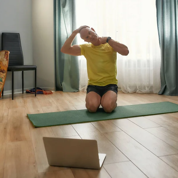Розігрівання вправ. Зрілий персональний тренер в спортивному одязі, дивлячись на ноутбук під час онлайн-тренувань вдома, показує вправи на йога килимок — стокове фото