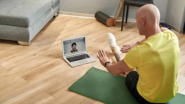 Вид спереду зрілого інструктора з фітнесу в спортивному одязі, що сидить на йога килимі вдома і проводить онлайн-тренування на ноутбуці, пояснюючи техніку тренування — стокове фото