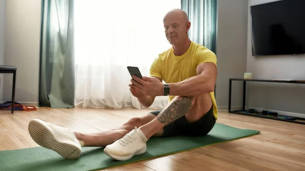 Інструктор з фітнесу вдома. Сильний атлетичний зрілий чоловік у спортивному одязі, використовуючи свій смартфон, сидячи на килимку йоги вдома, відпочиває після тренування — стокове фото