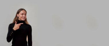 Siyah giyinmiş, sağ taraftaki bir şeyi işaret eden beyaz bir kadın gri arka planda yalnız poz veriyor.