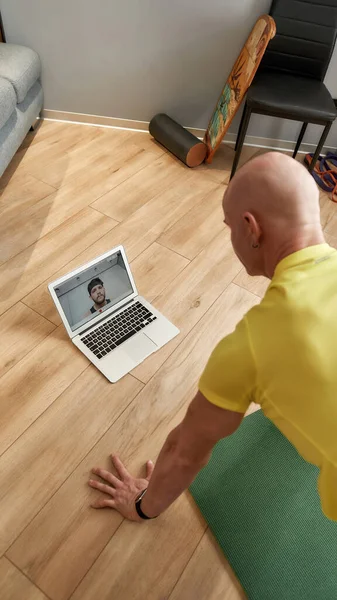 Проведення онлайн-тренінгів. Зрілий чоловік фітнес-інструктор в спортивному одязі робить поштовхи до йоги мат вдома і дивиться на ноутбук, маючи віртуальний фітнес-клас — стокове фото
