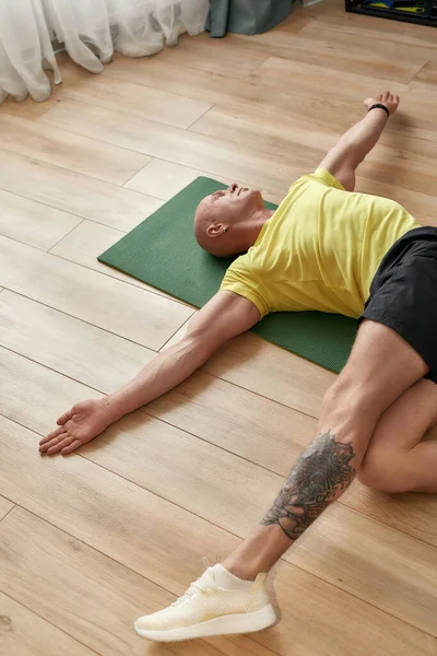 Yorgun sporcu bir adam gündüz vakti oturma odasında bir paspasın üzerinde dinlenirken evde yoga yapıyor.