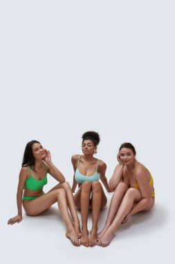 Renkli iç çamaşırlı üç genç kadının poz verirken, beyaz arka planda izole bir şekilde yerde otururken kameraya baktığı uzun bir çekim.