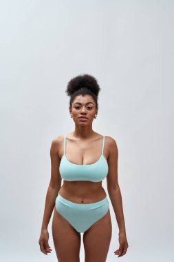 Mavi iç çamaşırı giyen güzel, seksi, genç, melez bir kadın. Işık arkaplanı üzerinde izole bir şekilde poz verirken kameraya bakıyor.