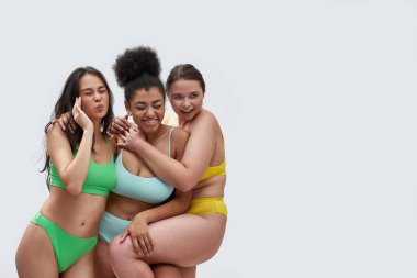 Renkli iç çamaşırlı üç genç kadın mutlu görünüyor, beyaz arka planda birlikte poz verirken eğleniyorlar.