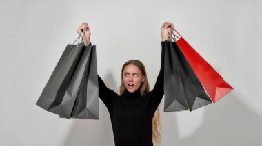 Surprised young caucasian woman wearing black clothes looking away, posing with arms raised holding shopping bags, standing isolated over light gray background