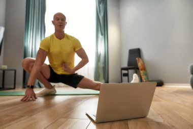 Virtual fitness class. Mature personal trainer in sportswear exercising and looking at laptop while conducting online training at home
