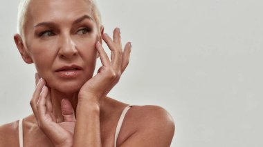 Anti wrinkle cosmetic products. Attractive caucasian mature woman in underwear touching her face and looking away while posing against light background