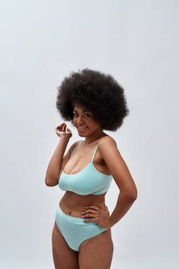 Playful curvy young female model with afro hair style wearing blue underwear smiling at camera, posing isolated over light background