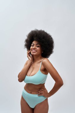 Adorable curvy young female model with afro hair style wearing blue underwear smiling at camera, posing isolated over light background