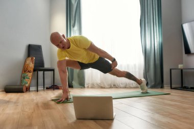 Mature personal trainer in sportswear looking at laptop while conducting online training at home, showing stretching exercises on yoga mat