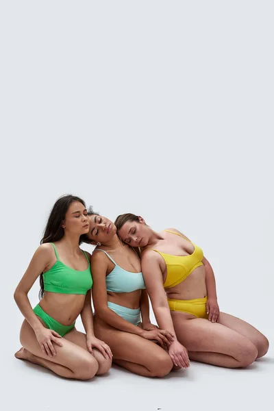 Full length shot of three diverse young women in colorful underwear posing together, leaning on each other, sitting on knees with eyes closed isolated over white background