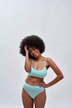 Heyecanlı, kıvırcık saçlı, mavi iç çamaşırı giyen, kameraya gülümseyen, arka planda izole bir şekilde poz veren genç bir model.