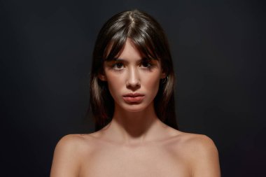 Büyük kahverengi gözlü ve renkli makyajlı çıplak esmer bir kadının güzel portresi. Kameraya bakıyor, siyah arka planda izole bir şekilde poz veriyor.