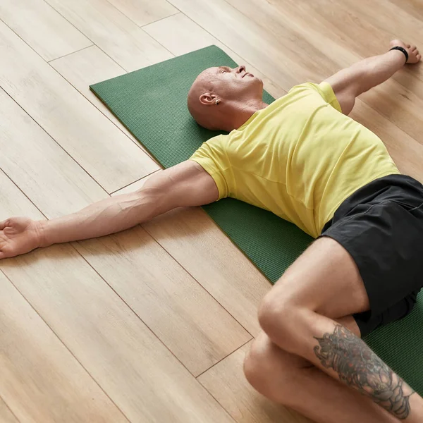 Yorgun sporcu adam evde yoga yapıyor, gündüz vakti oturma odasında bir paspasın üzerinde dinleniyor.