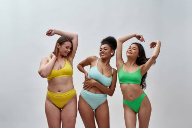 Farklı vücut şekillerine sahip güzel genç kadınlar renkli iç çamaşırları giyerek stüdyoda ışık arka planında birlikte poz verirken eğleniyorlar.