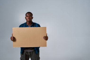 Ciddi bir Afro-Amerikan vatandaşı. Günlük kıyafetleriyle kameraya bakıyor. Önünde boş karton pankart ilanı tutuyor.